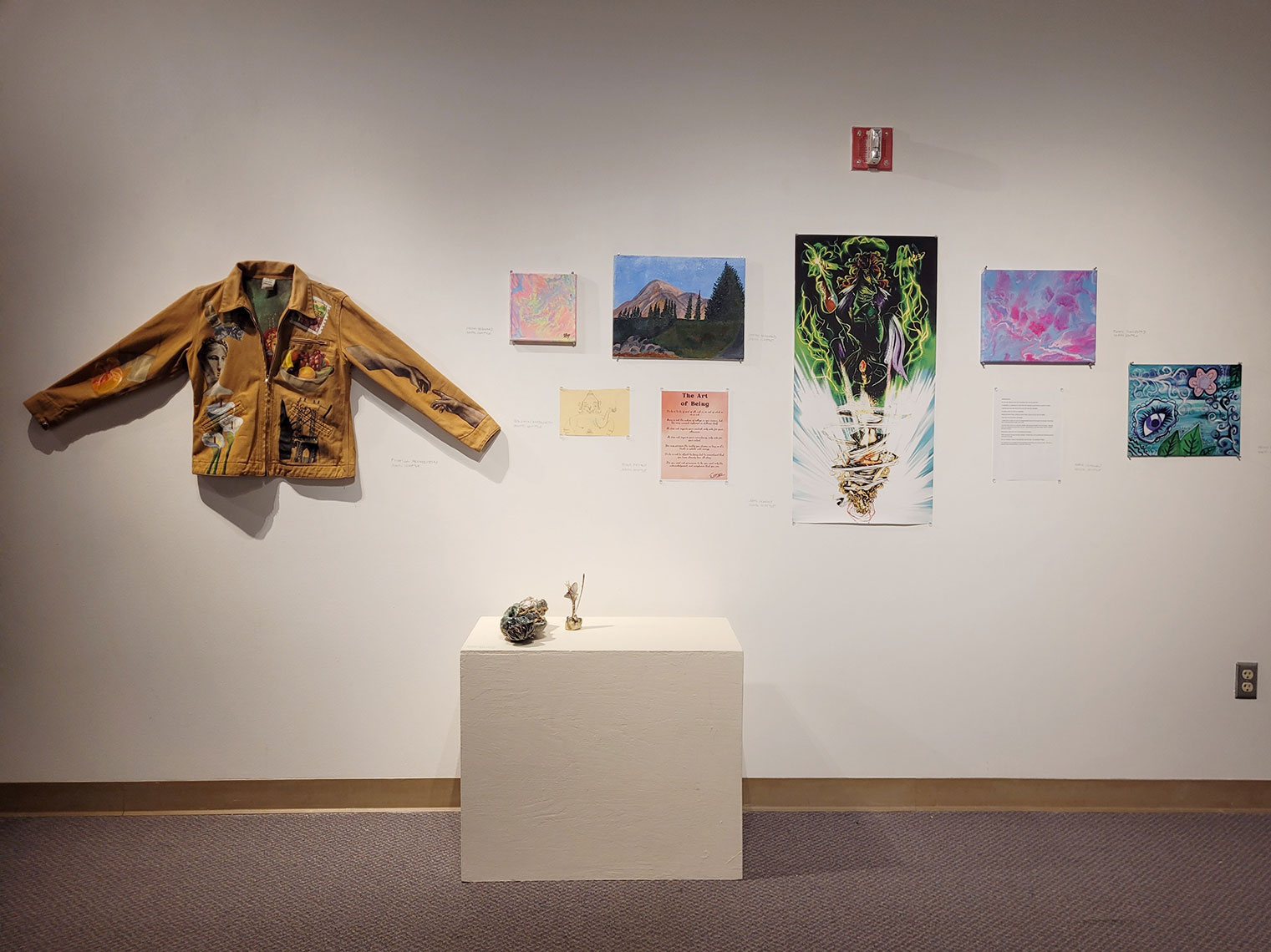 A photograph of an art installation with paintings and drawings on the wall, and a painted jacket. In front of the installation is a low pedestal showcasing ceramic sculptures.